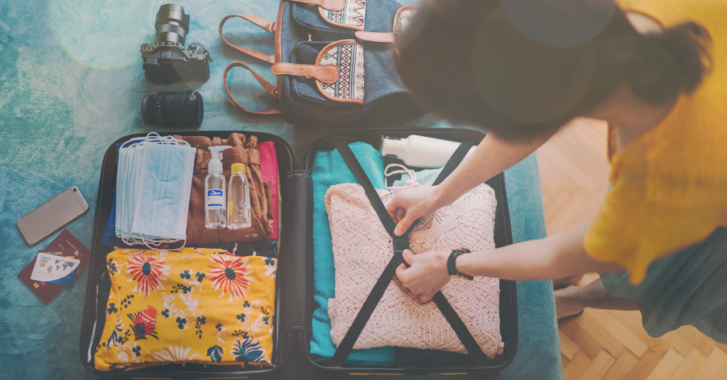 Various items on a bed and clothes next to a woman packing a organized suitcase