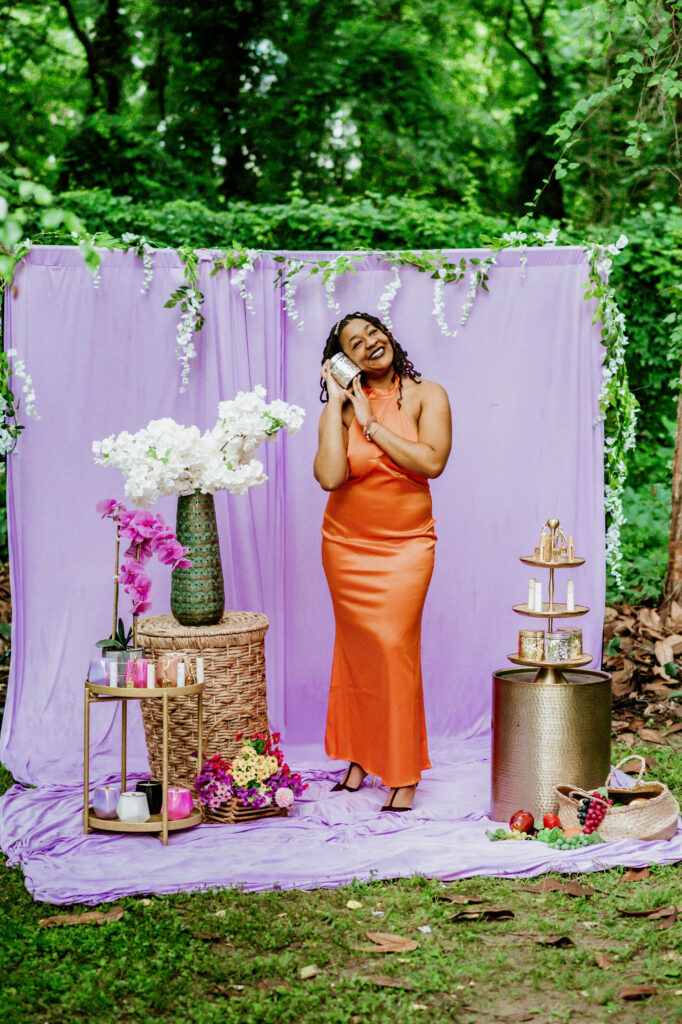 Beautiful black woman in a orange silk dress, surrounded by greenery and a purple backdrop. She is accessorized and highlighting candles