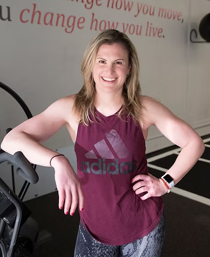 A white woman with blonde hair in a purple shirt and workout pants, posing in a strong stance in a gym.